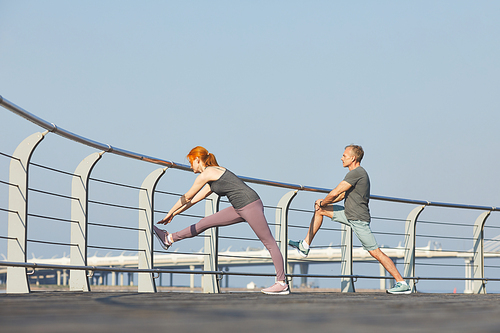 Sporty modern mature couple using railing on embankment while stretching leg muscles before running