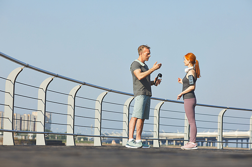 Redhead mature woman with smartphone on arm planning outdoor training with fitness instructor while they talking on embankment