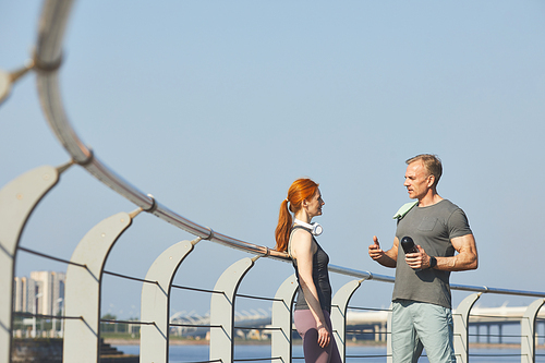 Professional fitness coach with water bottle standing on embankment and discussing training with redhead woman