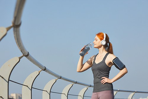 Energetic redhead mature woman in wireless headphones drinking water after training on riverside