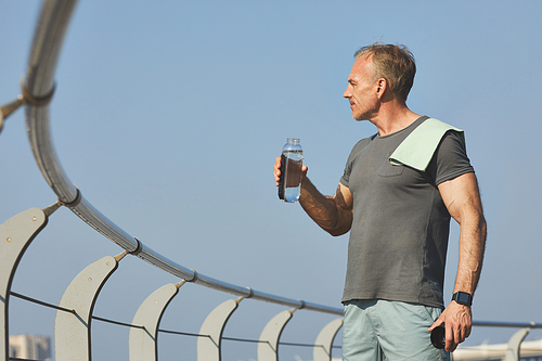 Handsome muscle-bound man with towel on shoulder contemplating riverside while drinking water after training