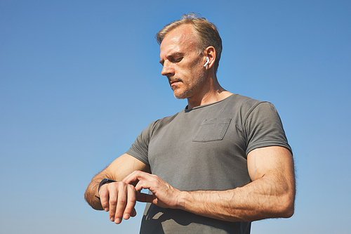 Confident handsome mature man in ear buds standing against blue sky and taking pulse with smartwatch