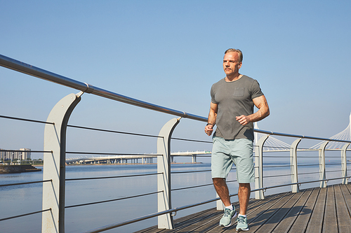 Athletic mature man in shorts jogging on city embankment in morning, healthy lifestyle concept