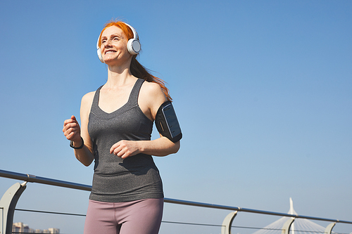 Below view of smiling energetic redhead woman listening to music in wireless headphones while running in city