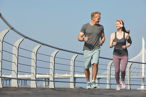 Positive active mature couple in headphones looking at each other while jogging together on embankment