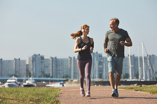 Sporty mature couple in headphones smiling at each other while enjoying jogging together in city