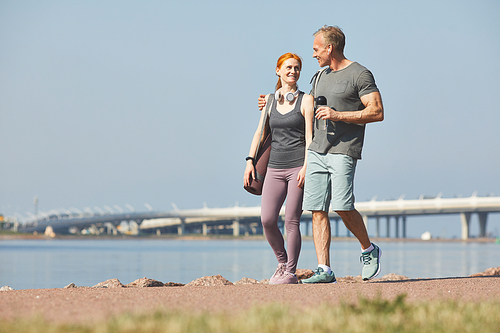 Smiling mature man with water bottle embracing wife while walking with her over riverside after training