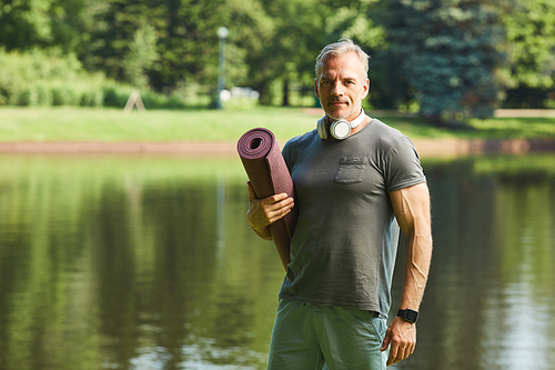 Portrait of content muscular mature man with headphones around neck holding exercise mat against park lake