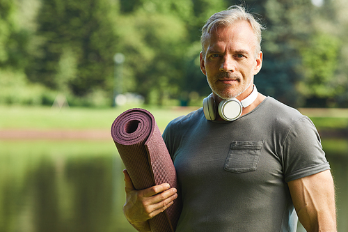 Portrait of handsome mature man with wireless headphones around neck holding rolled fitness mat in city park