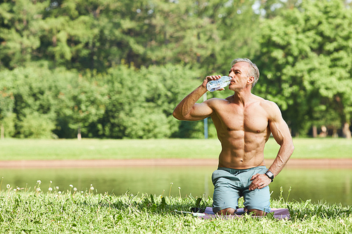 Handsome muscular man with abs on belly sitting on mat in park and drinking water from bottle