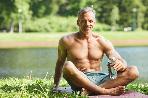 Portrait of smiling handsome mature sportsman sitting with crossed legs on mat and drinking water while resting at riverside