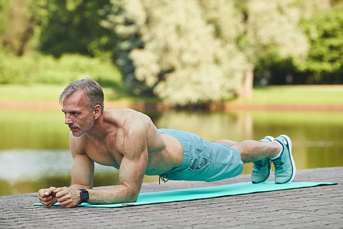 Concentrated muscle bound mature man with shirt doing plank on exercise mat near river