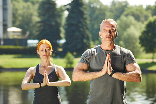 Calm mature couple with closed eyes keeping hands in Namaste and meditating together at river in city park