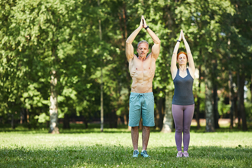 Serious sporty mature couple standing with raised hands in Namaste and doing breath exercise in summer park