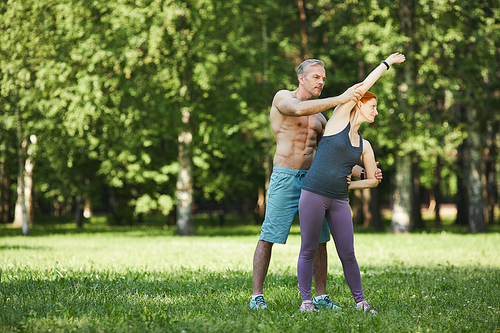 Personal shirtless fitness instructor assisting woman to do side bend in park during coronavirus restrictions