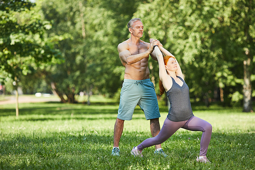 Professional muscle-bound trainer adjusting yoga pose of woman while they practicing in summer park