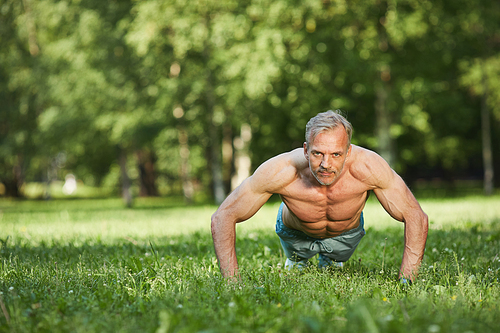 Portrait of strong purposeful mature man with muscular arms doing push-ups from ground in park