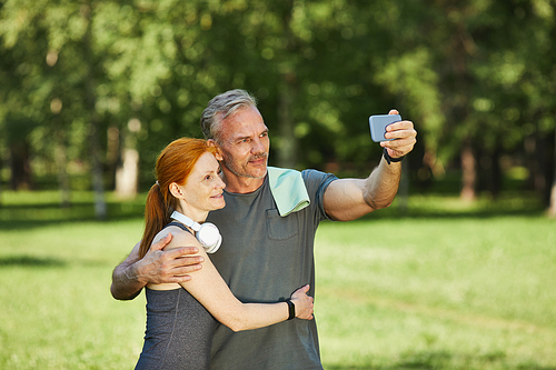 Handsome mature sportsman with towel on shoulder embracing wife and photographing with her after training in park