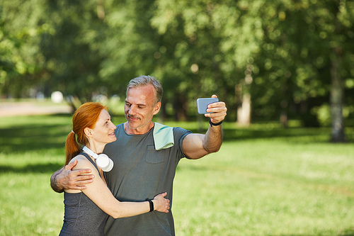 Loving mature couple looking at each other and embracing while posing for selfie in park after sports training