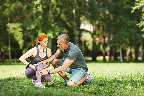 Careful mature fitness trainer checking knee of woman while she feeling pain after exercise in park