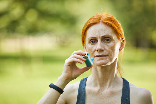 Portrait of serious freckled mature woman using inhaler to prevent asthma attack after training in park