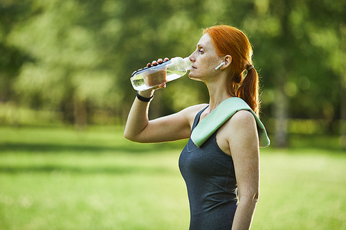 Side view of mature redhead fitness woman with towel on shoulder drinking water in park after exercising