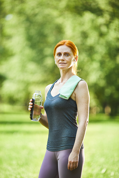 Portrait of smiling confident redhead mature woman with towel on shoulder holding bottle of water in summer park