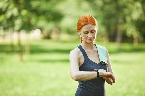 Attractive redhead mature woman using fitness tracker while checking pulse after outdoor training in park