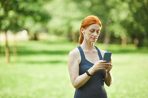 Redhead mature woman in wireless ear buds standing in park and choosing music for fitness using smartphone