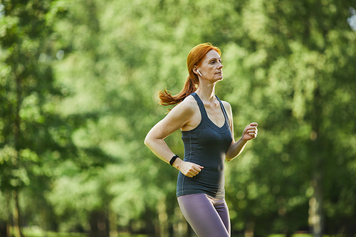 Serious mature redhead woman in wireless earphones wearing fitness tracker running in green park