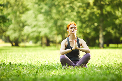 Serene redhead mature woman sitting on grass and holding hands in Namaste while meditating in empty park