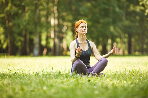 Serene relaxed mature redhead woman sitting with crossed legs on grass and meditating with closed eyes in park