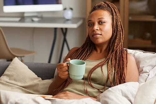 Portrait of young woman with beautiful hairstyle looking at camera while resting on sofa and drinking coffee