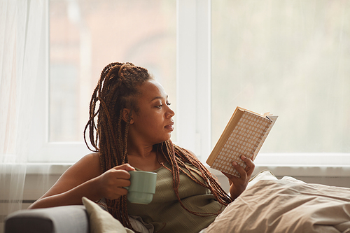 African young woman lying on sofa drinking coffee and reading an interesting book at home