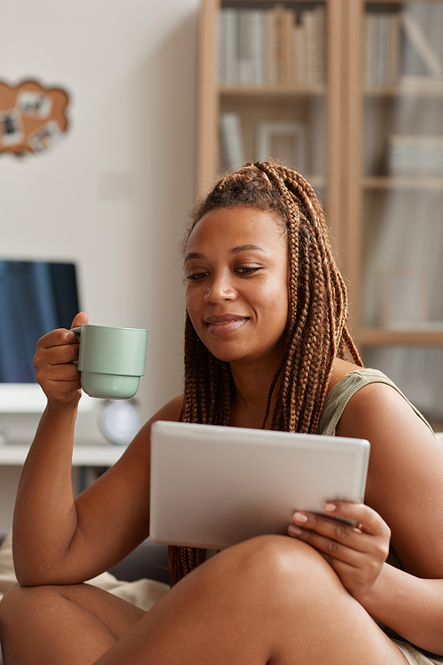 African young woman sitting on sofa with cup of coffee and using digital tablet at home