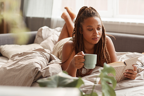 Young woman lying in bed drinking coffee and reading a book in the morning she enjoying her weekends