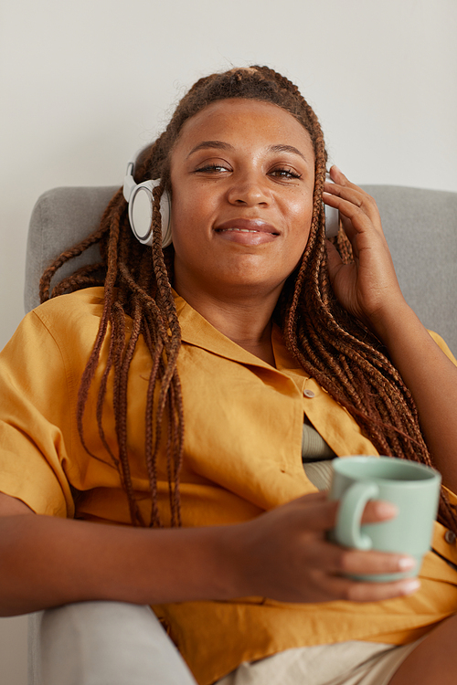 Portrait of African woman in headphones smiling at camera while resting on armchair with cup of coffee