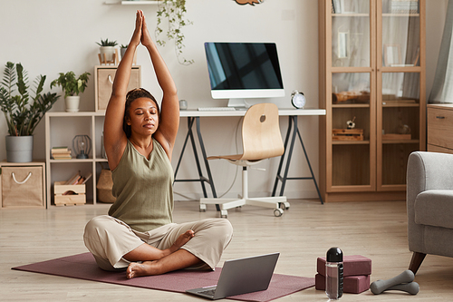 Young woman sitting on exercise mat in front of laptop and doing yoga at home