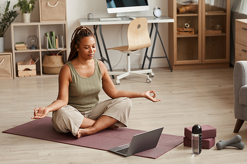 Young woman sitting in lotus position with laptop near her and doing yoga online in the room