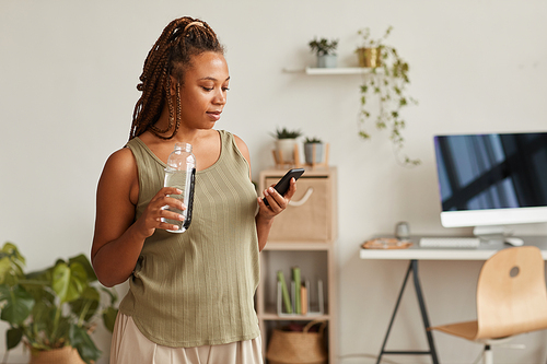 Young woman drinking water from the bottle and typing a message on her mobile phone she resting after sports training at home