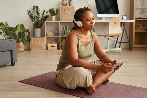 African woman in wireless headphones sitting on exercise mat with her eyes closed and listening to relaxing music using digital tablet
