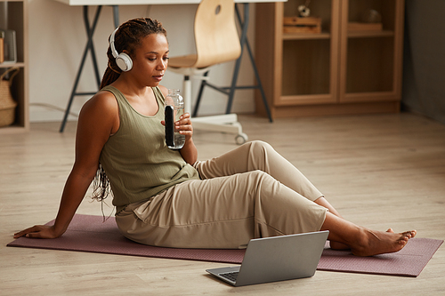 Young woman in headphones sitting on exercise mat drinking water and watching sport exercises online on laptop at home