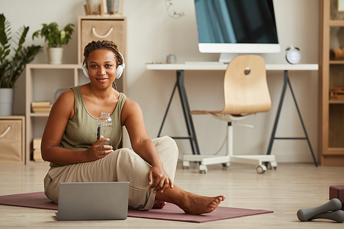Portrait of African young woman in headphones drinking water and looking at camera while sitting on exercise mat and resting after training
