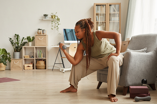 African woman sitting on armchair and training her hands with dumbbells at home
