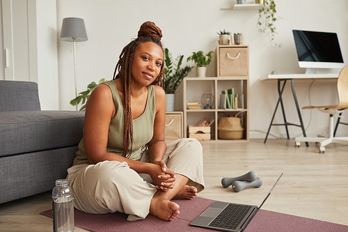 Portrait of African young woman with beautiful hairstyle sitting on exercise mat and using laptop during her sport training at home