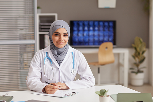 Portrait of young female doctor in white coat looking at camera while sitting at the table at office
