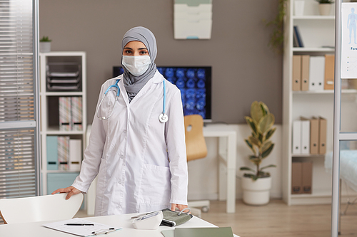 Portrait of muslim doctor in white coat and in protective mask looking at camera while working at clinic