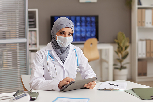 Portrait of muslim doctor in protective mask sitting at the table using digital tablet while working at hospital