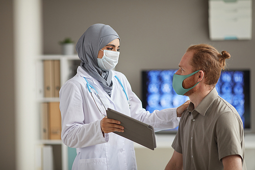 Muslim female doctor in mask holding digital tablet and discussing with patient his disease at hospital