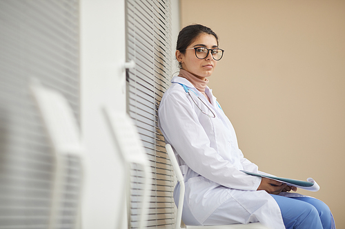 Portrait of young nurse in white coat looking at camera while sitting at the corridor of hospital
